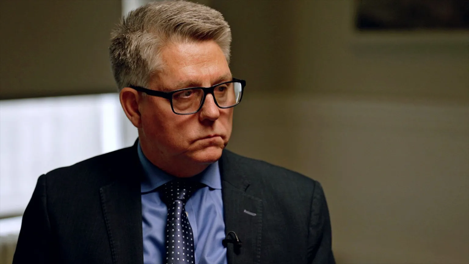 Adam Walker/BBC Kevin Hyland, a man with fair hair and dark-framed glasses, wearing a dark suit, blue shirt and navy tie, pictured in a low-lit, sparsely decorated room, with his face lit softly and the light from outside visible through the half-drawn blind.