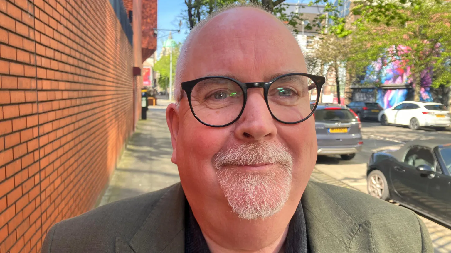 A man, who is bald and has grey stubble, wearing glasses and a dark green blazer. there is a red brick wall to his left and a row of parked cars and trees to his right. 