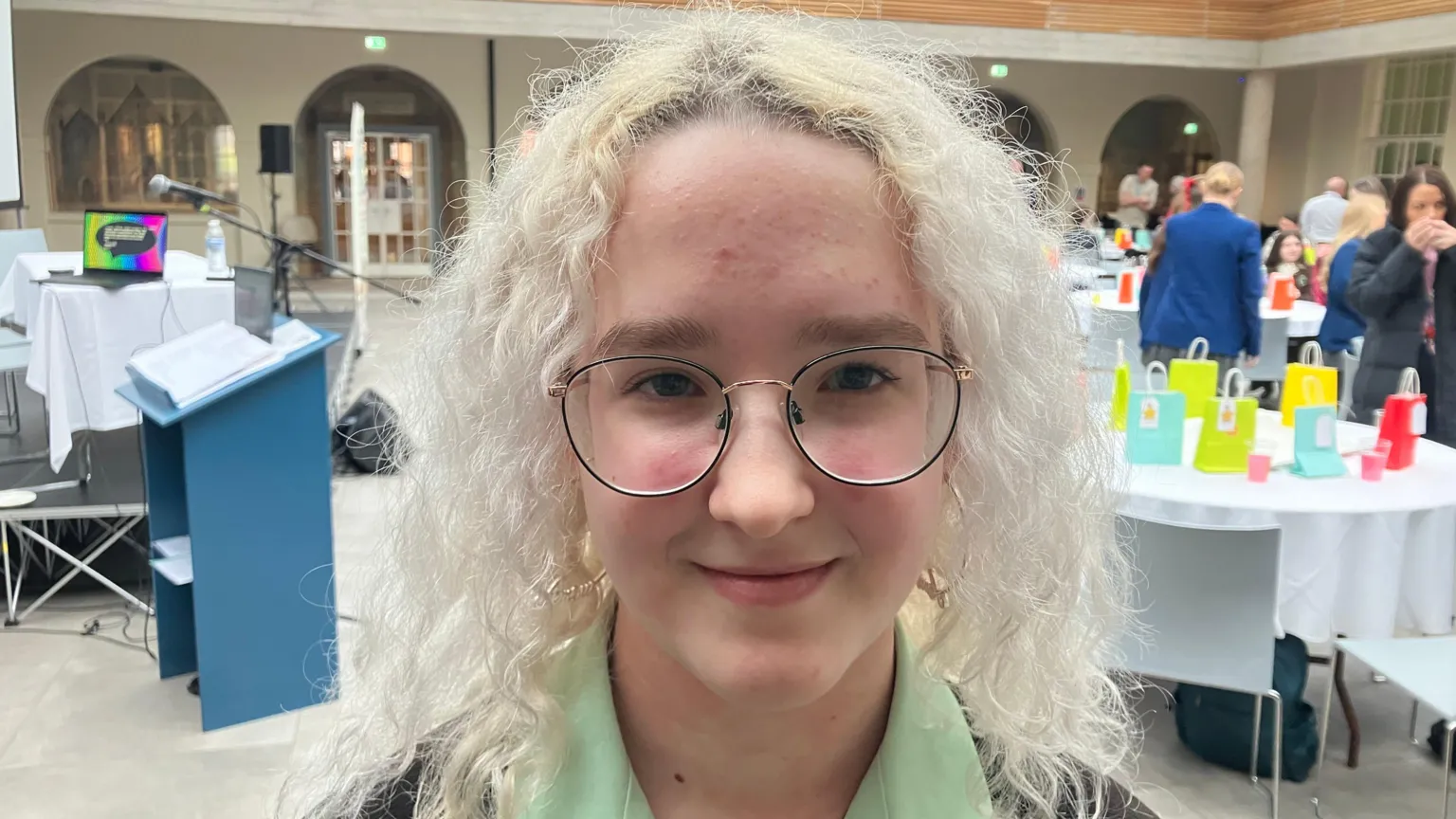 A teenage girl, wearing glasses, a green shirt and a brown jumper. She has blonde curly hair. 