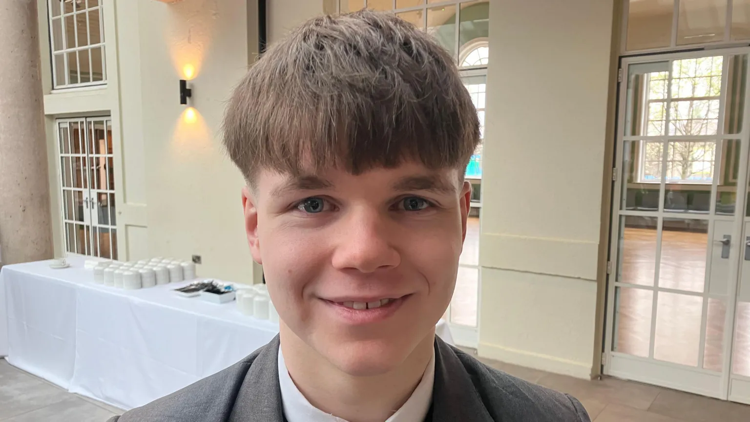 A teenage boy, wearing a white shirt and grey blazer. He has short brown hair. 