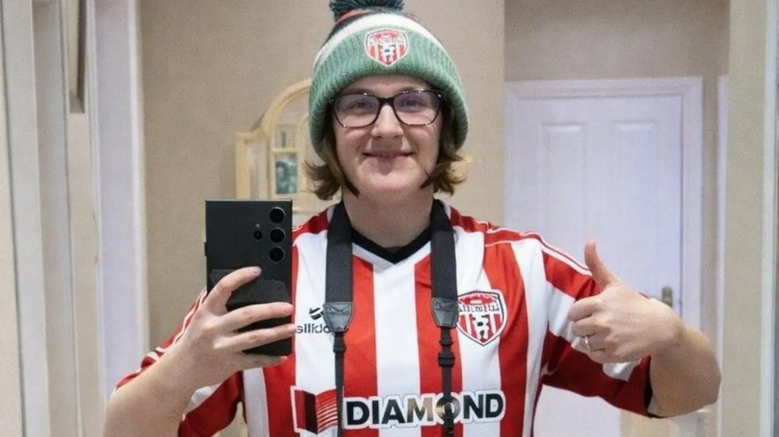 Claire McCarron Claire McCarron is wearing a red and white Derry City shirt. She is taking a mirror selfie and has a Derry City woolly hat on as well. She is holding a large black phone in her right hand and doing a thumbs up with her left hand. 