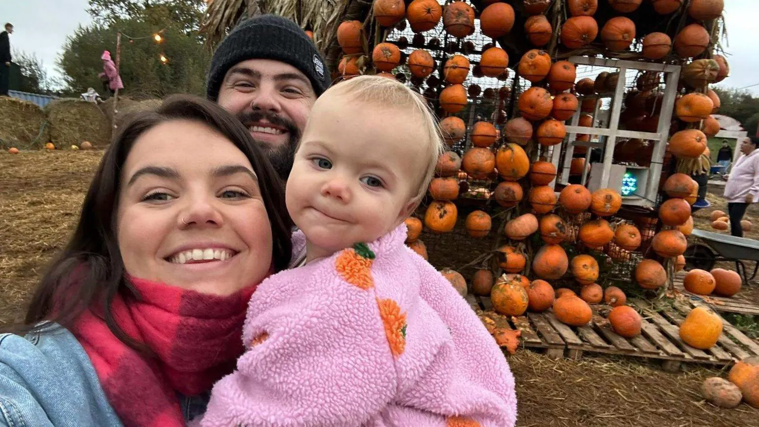 Gareth Williams A mum and dad in their thirties with a toddler in a pink fleece with pumpkins on it. It is around Halloween time with a display on pumpkins in the background in a muddy field. 