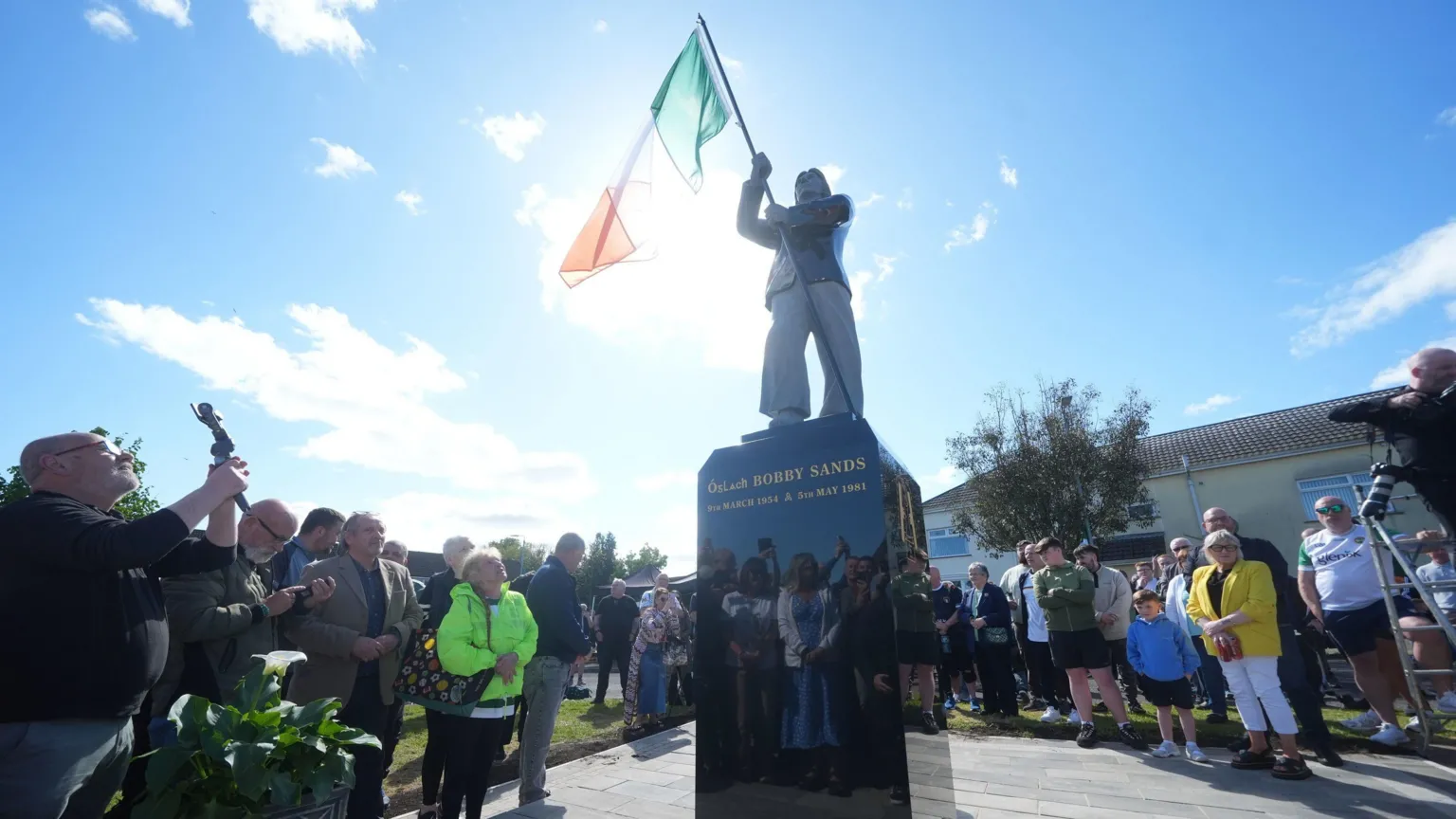  Members of the public attend the unveiling of the Bobby Sands statue. The statue is in the centre of the photo with crowds of people standing around it. It is a sunny day. 