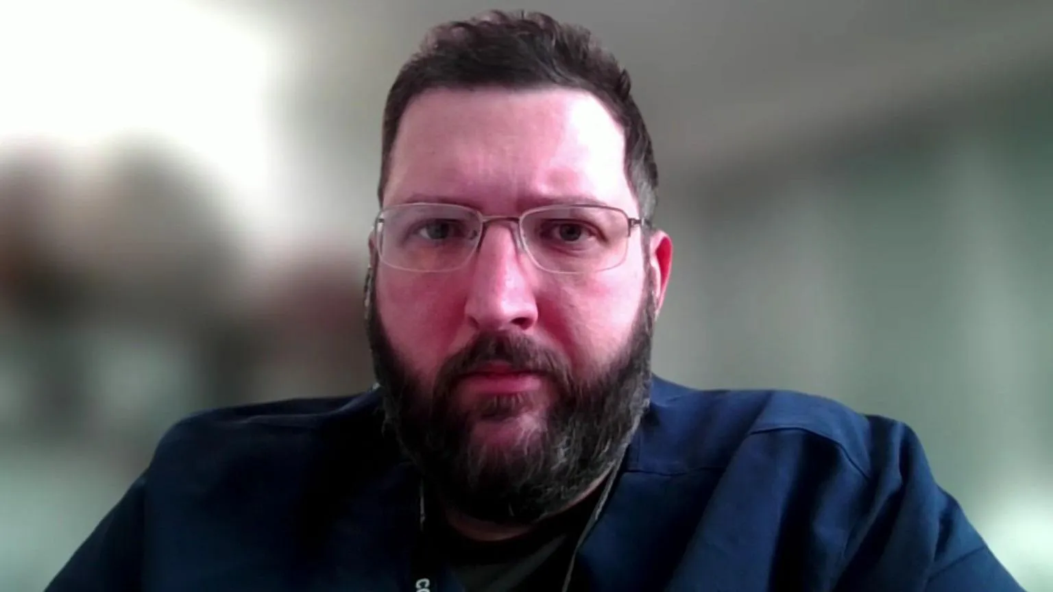 Michael Perry looks into the camera. He has short brown hair and a beard and wears glasses and is wearing a dark blue shirt. The background is blurred.