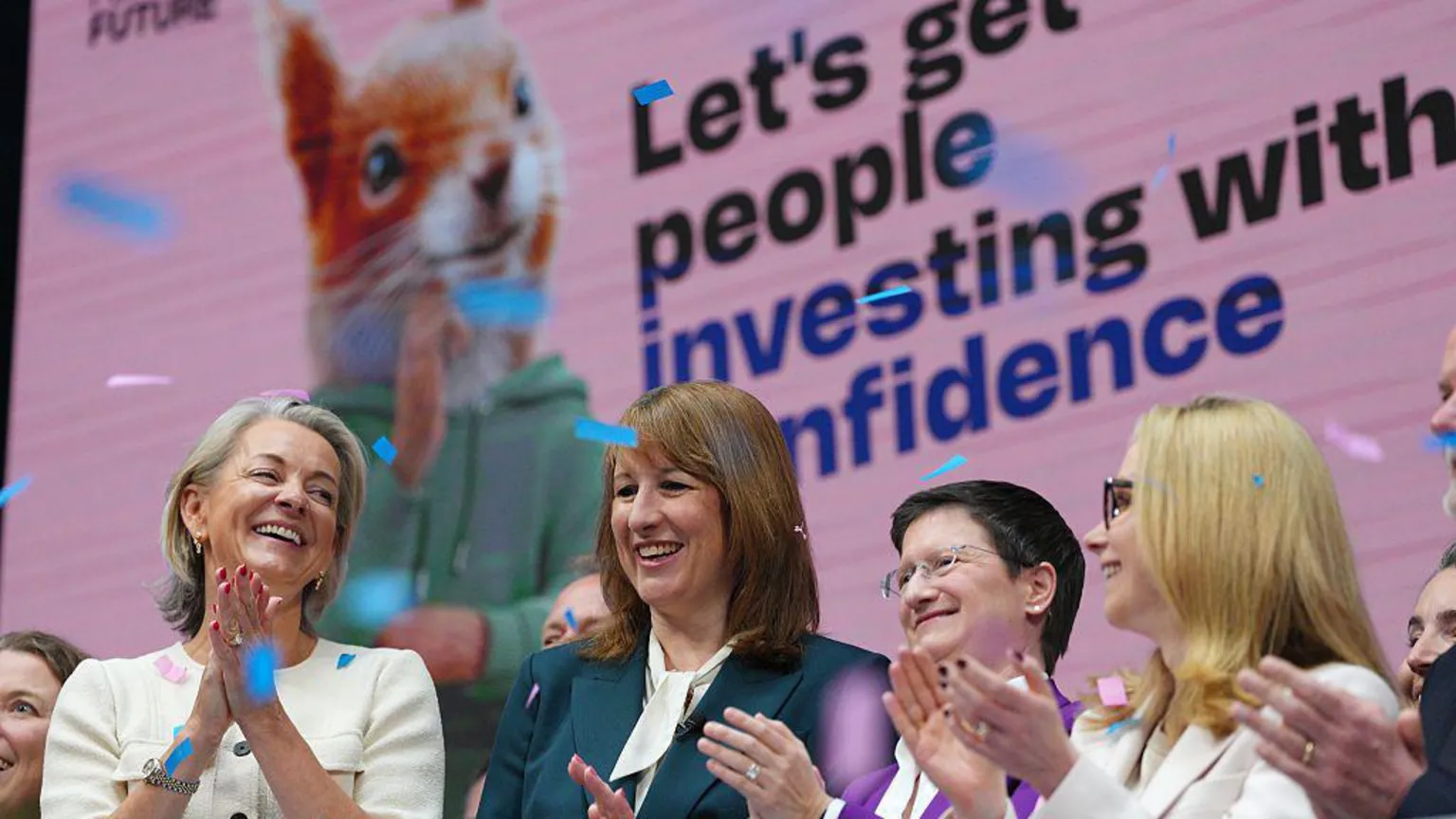  Chancellor Rachel Reeves, Julia Hoggett, CEO of London Stock Exchange, and Lucy Rigby, Economic Secretary to the Treasury, are seen at the launch the investment campaign, at the London Stock Exchange on April 23, 2026. They are clapping at the opening of the market with the advert on a large screen behind them.