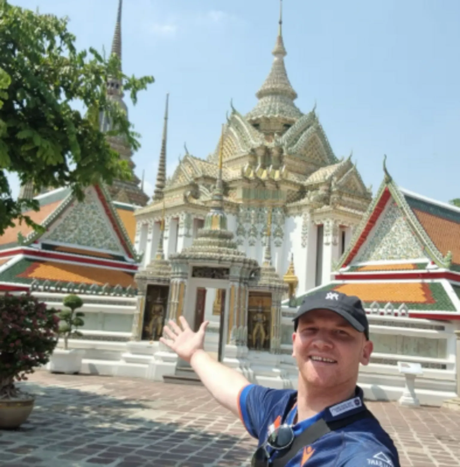 Rory McColl standing in front of Thai architecture, gesturing behind him. He is smiling and wearing a blue polo shirt and a black cap.