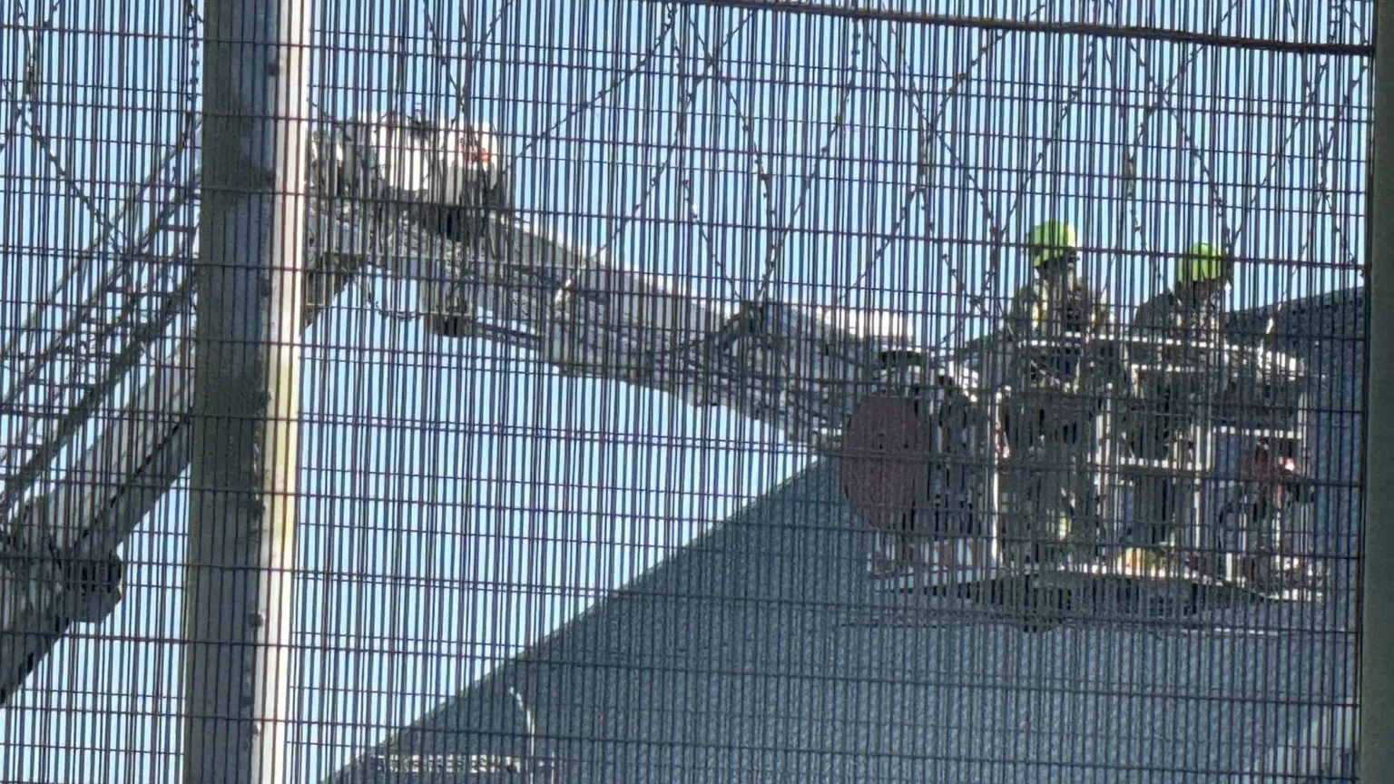 Jamie Niblock/BBC Firefighters in the cage of an aerial ladder platform. There appear to be two of them. They are approaching a roof. The shot has been taken through a wire fence at Highpoint Prison