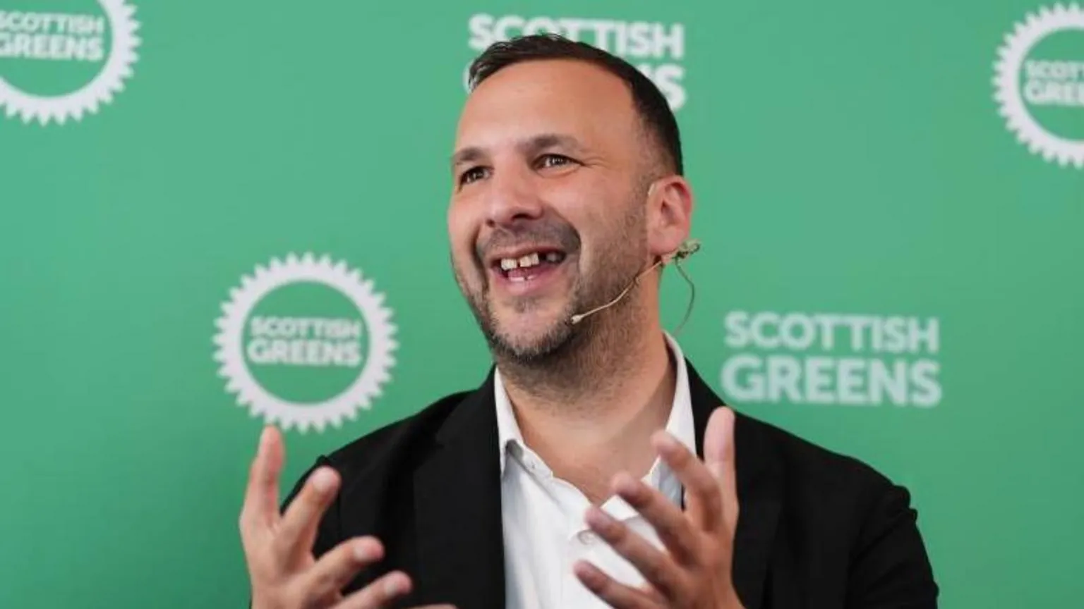  Zack Polanski, a man with short brown hair wearing a white shirt and black suit jacket. He is mid speech wearing a microphone. He has his hands gesturing in front. Behind him is a green background with the Scottish Greens logo on it.