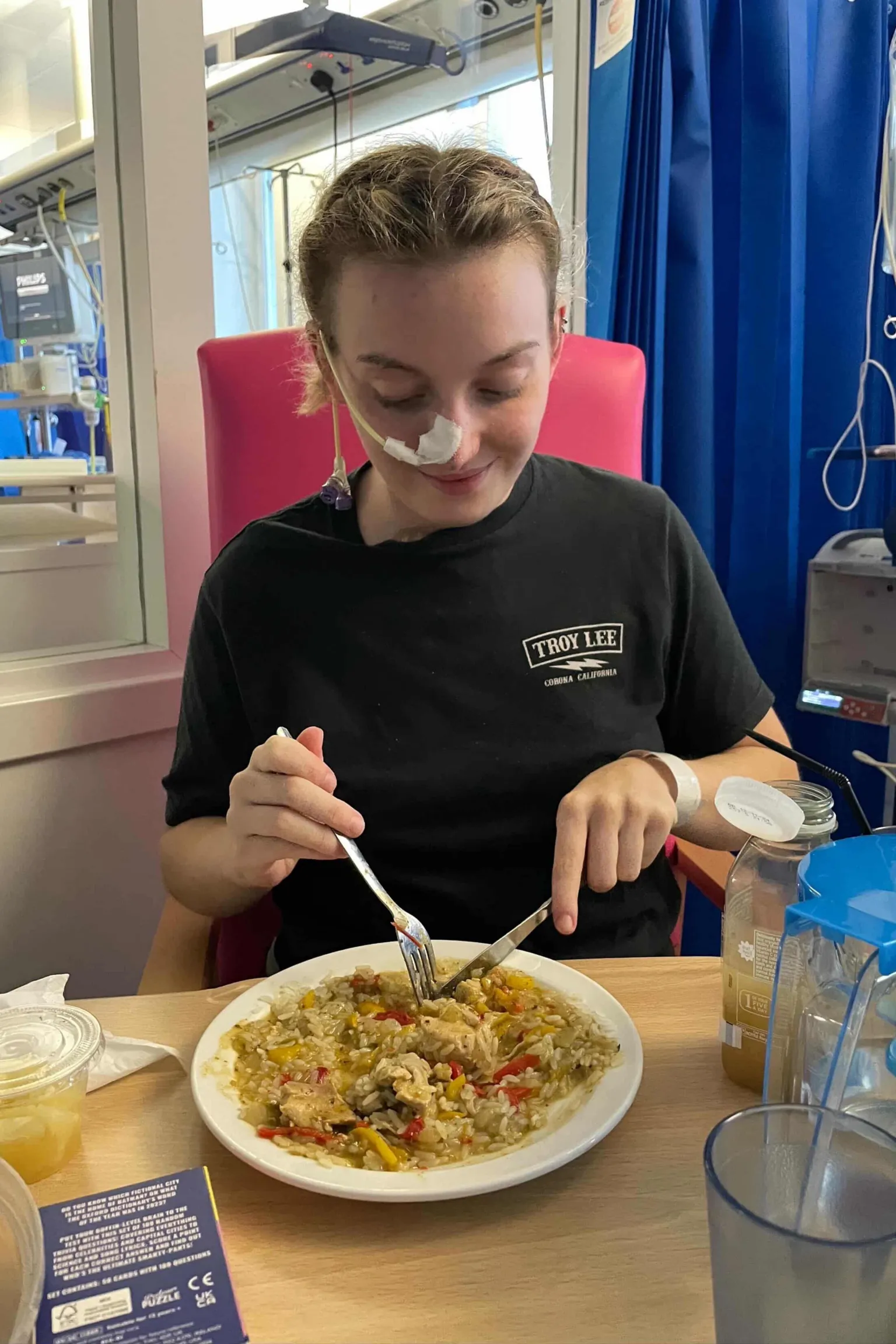 Katie Bain Katie with a tube attached to hr nose is sitting in a hospital room, she is bearing black t-shirt and eating a meal.