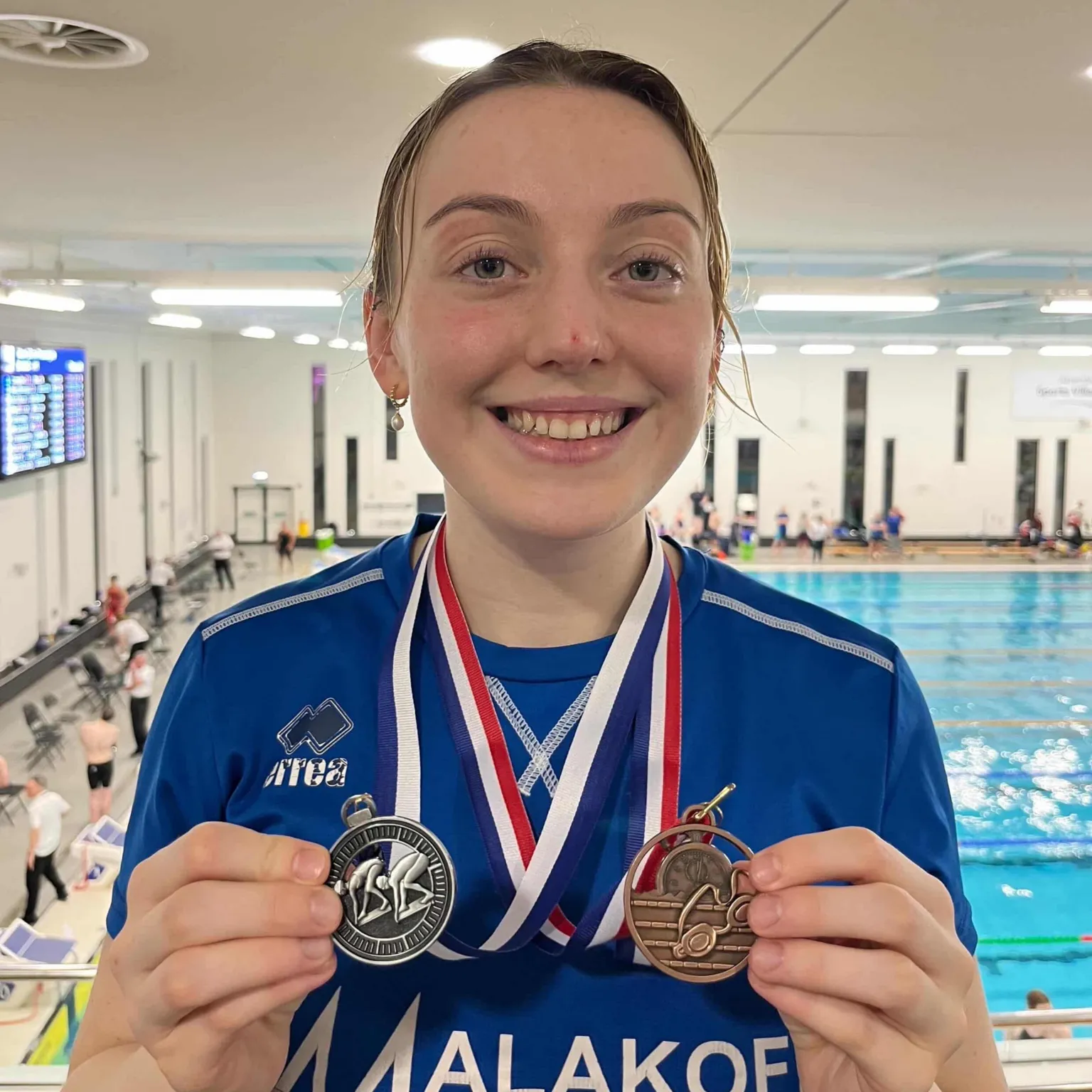 Katie Bain Katie is in the indoors swimming pull, the pool itself id behind her, she is wearing navy blue t-shirt and is holding silver medal in one hand and bronze in the other