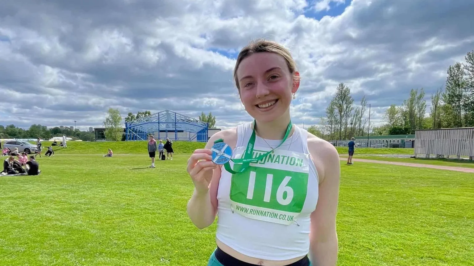 Katie Bain Katie is standing in the middle of the shot, she wears a white sleeveless top with her race number sticker, from when she ran 3 miles (5 km) in April 2025. Green lawn is behind her, it is bright sunny day. 