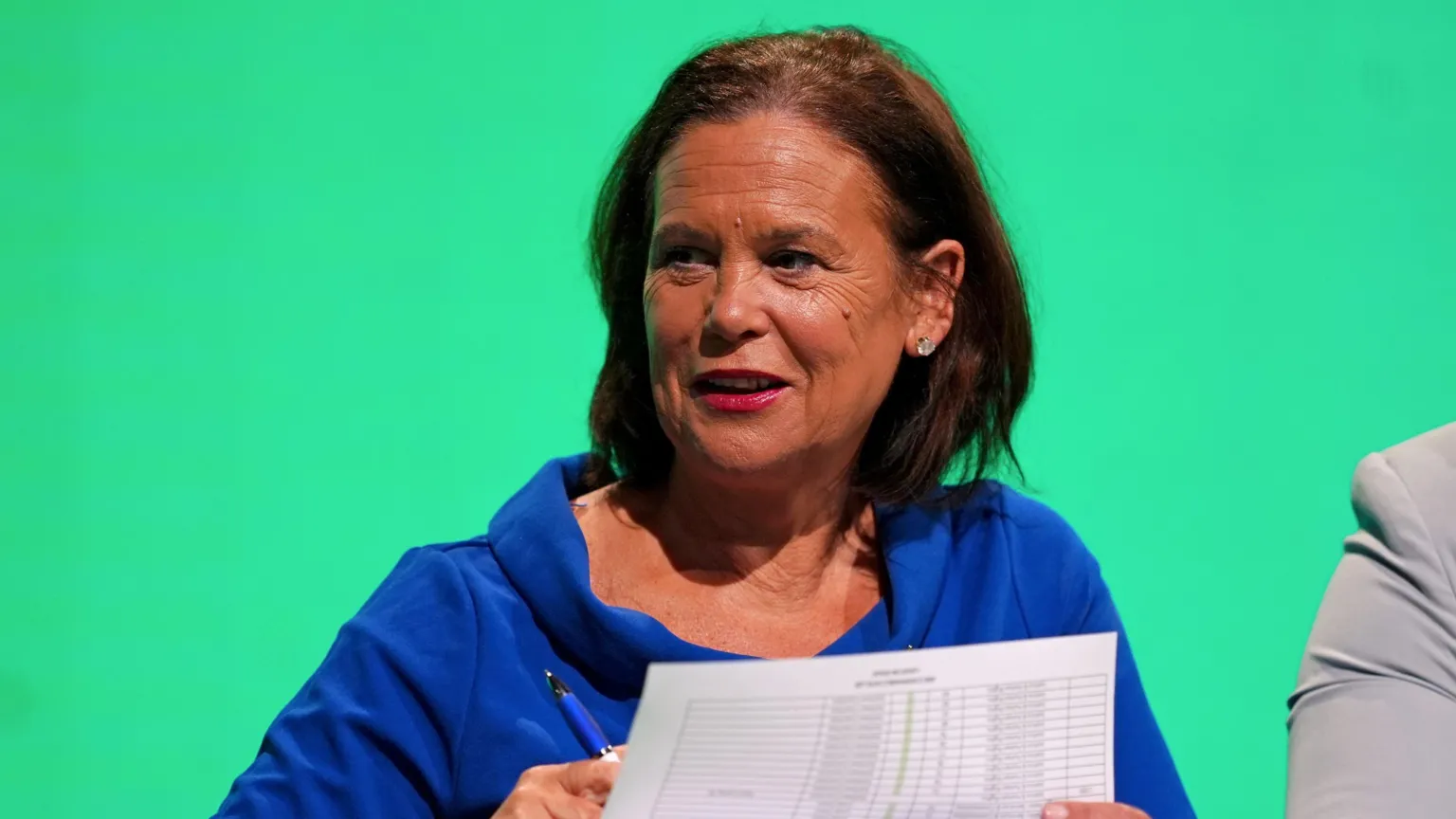  Sinn Féin leader Mary Lou McDonald at the Sinn Féin Ard Fheis. She has shoulder length brown hair and is wearing a bright blue top. The background behind her is green.