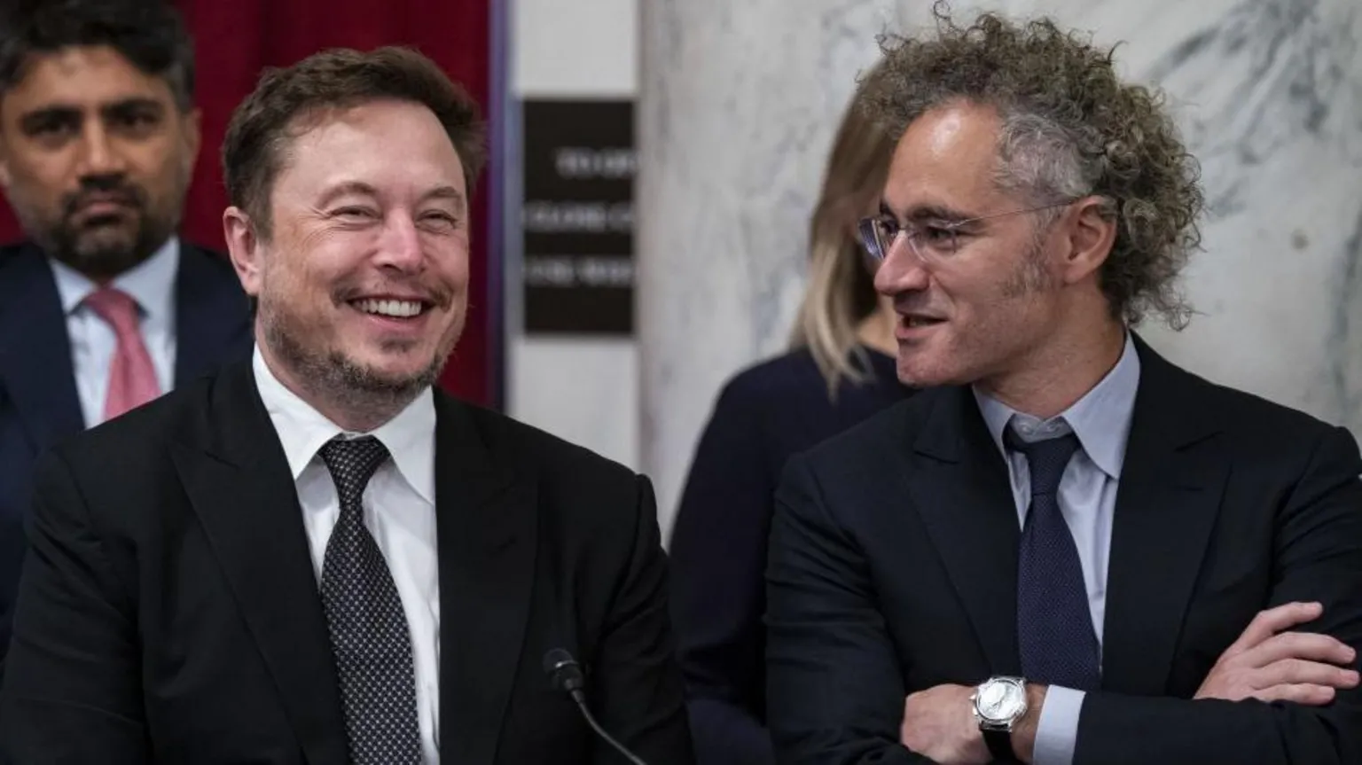  Elon Musk and Alex Karp sitting next to each other. Both wear dark suits and ties and are laughing.