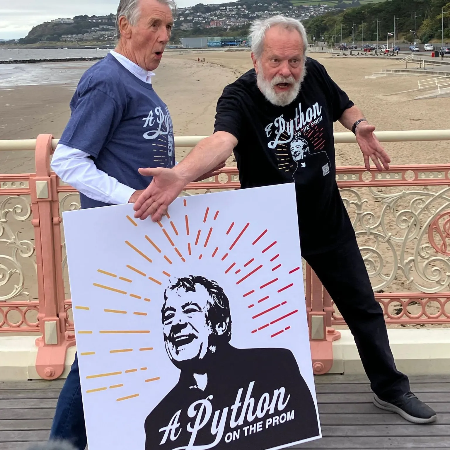 Sir Michael Palin and Terry Gilliam strike silly poses on Colwyn Bay pier while holding a poster featuring Terry Jones and advertising the Python on the Prom campaign