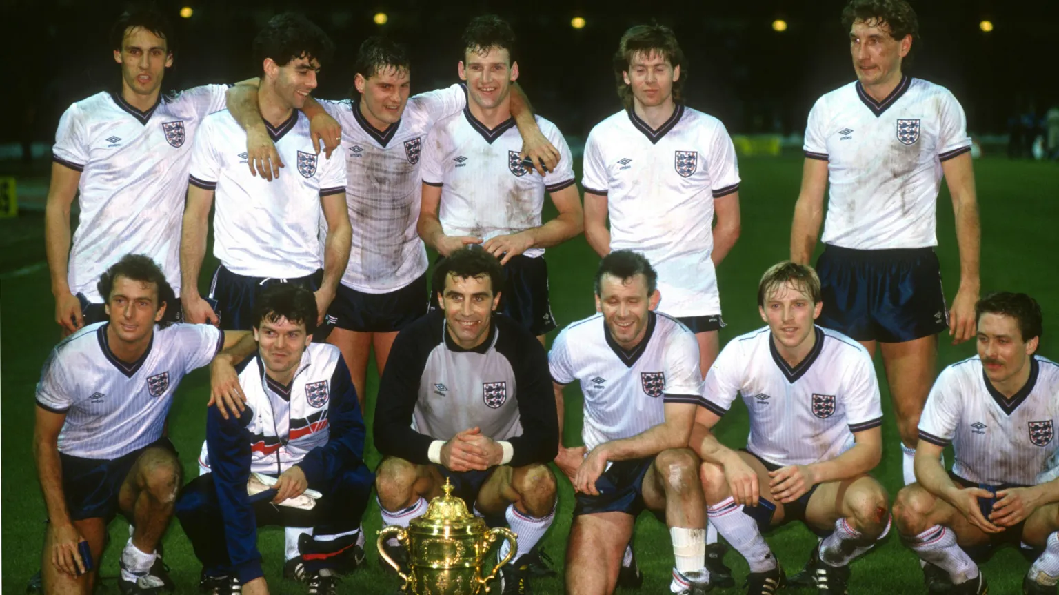 Offside via An England team photo, with 13 men dressed in white shirts and blue shorts lined up in two rows on a green pitch following a match. Players in the back row have their arms around one another, while those in the front row are kneeling behind a large gold trophy.