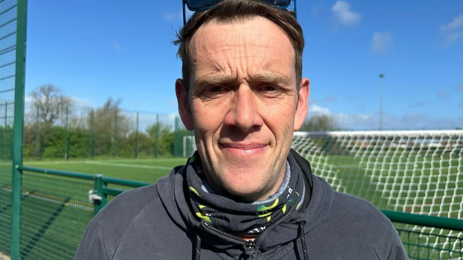 A man wearing a neck scarf and a zip up hoodie is standing in front of a green football training pitch with goals visible in the background. The sky is blue.