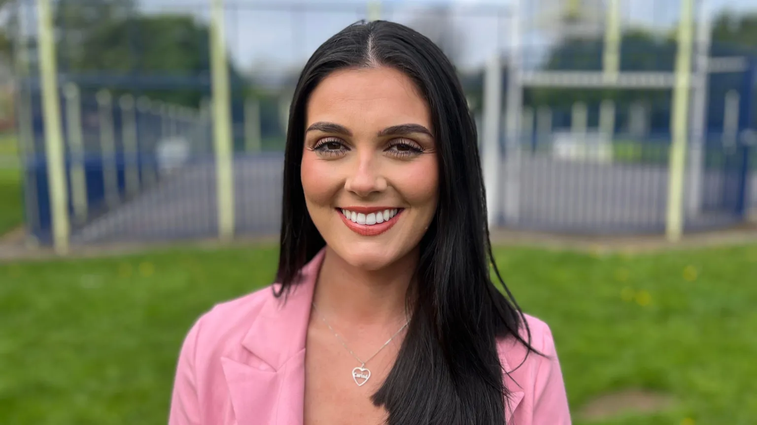 A woman with long dark hair, wearing a pink jacket and a silver heart necklace, standing in a park with a basketball court in the background.