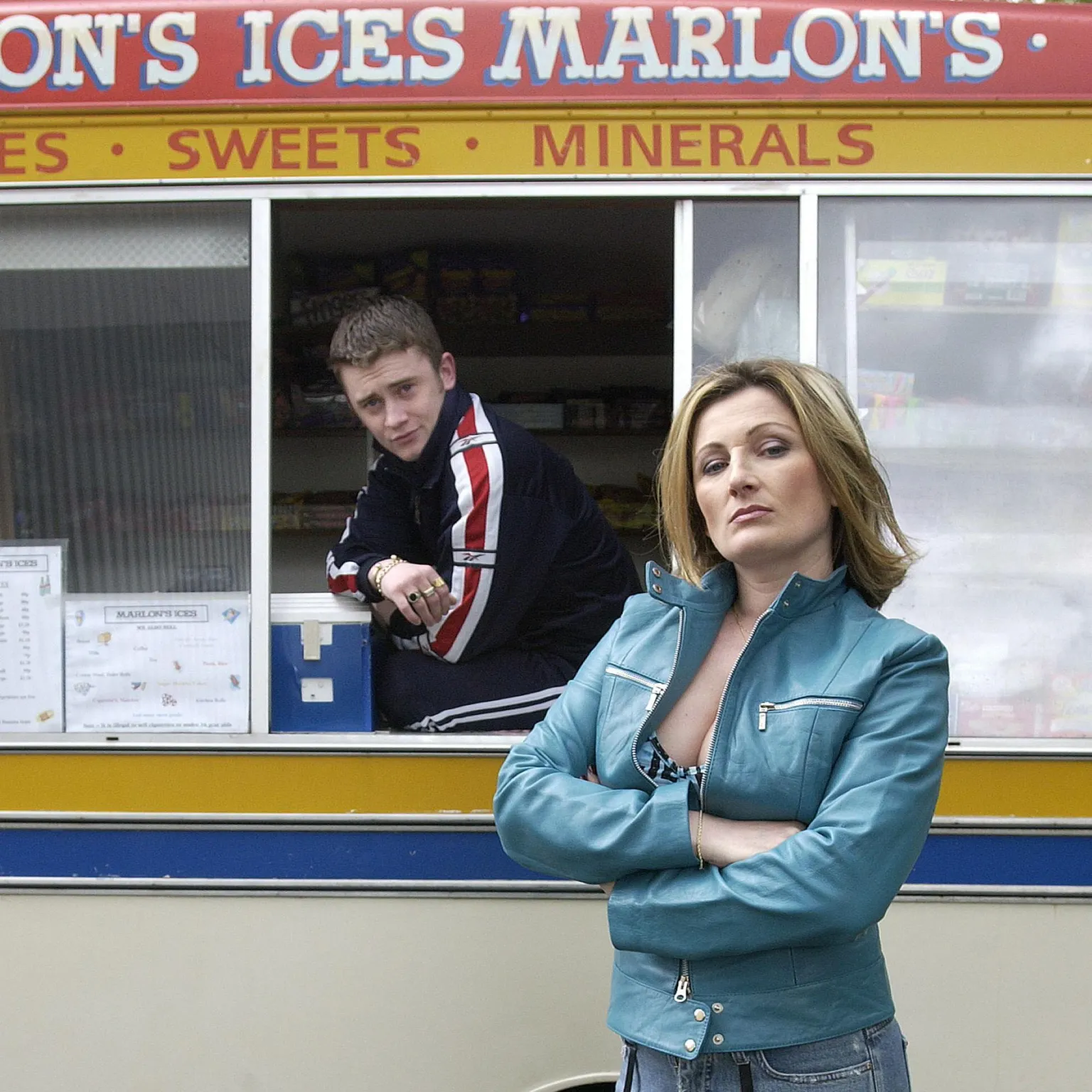BBC Studios Drama A person stands in the foreground with arms folded, wearing a turquoise jacket, patterned jeans, and holding a leopard‑print bag. Behind them is a cream-and-yellow ice cream van with signage reading “Marlon’s Ices” and “Sweets • Minerals.” Another person leans out of the serving window of the van, suggesting a casual street setting on a wet roadway.