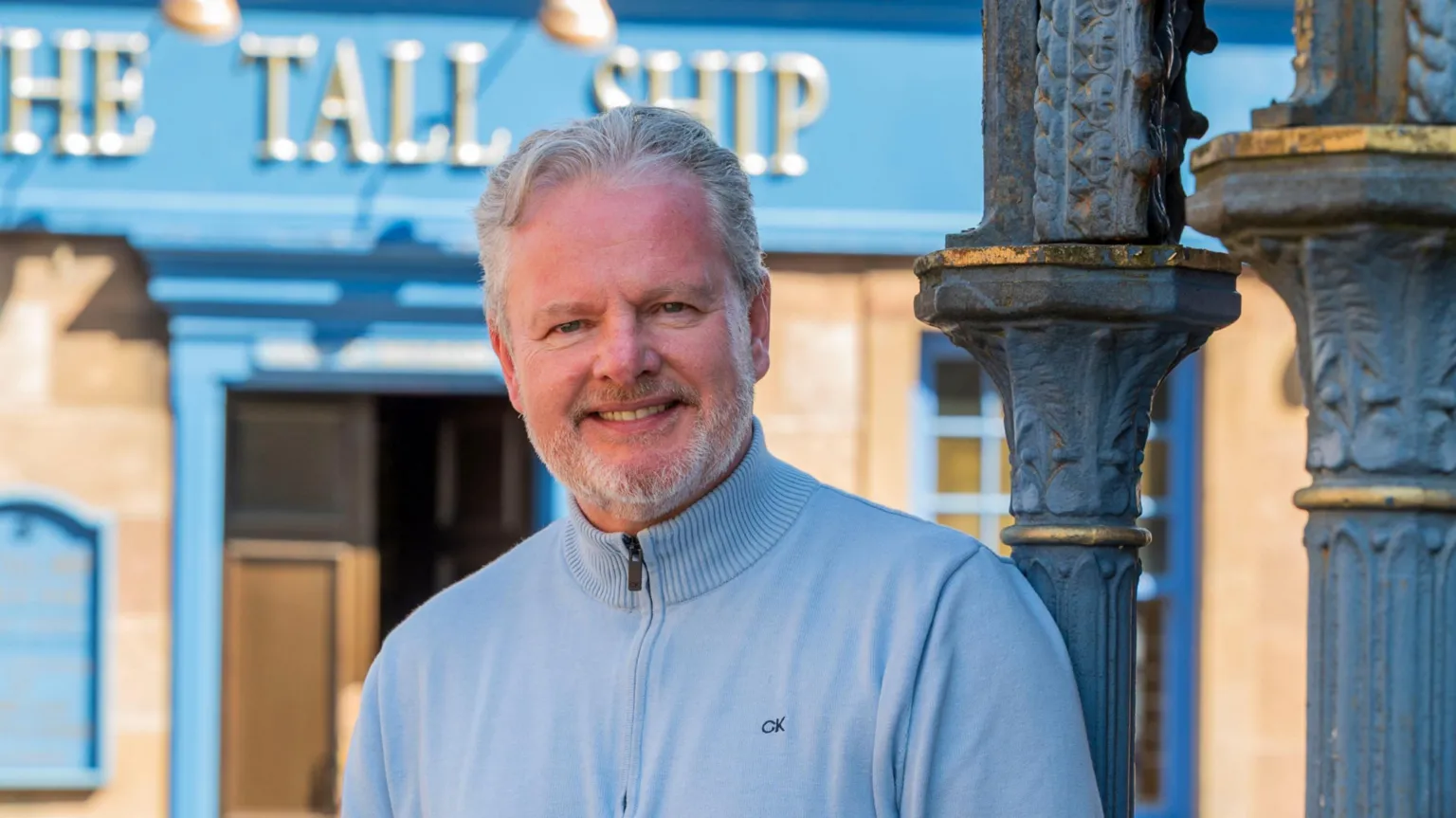 BBC Studios Drama A person stands outdoors beside a decorative cast‑iron column, wearing a light blue sweater and jeans. Behind them is a stone building with bright blue trim and a sign reading “The Tall Ship” above the doorway.