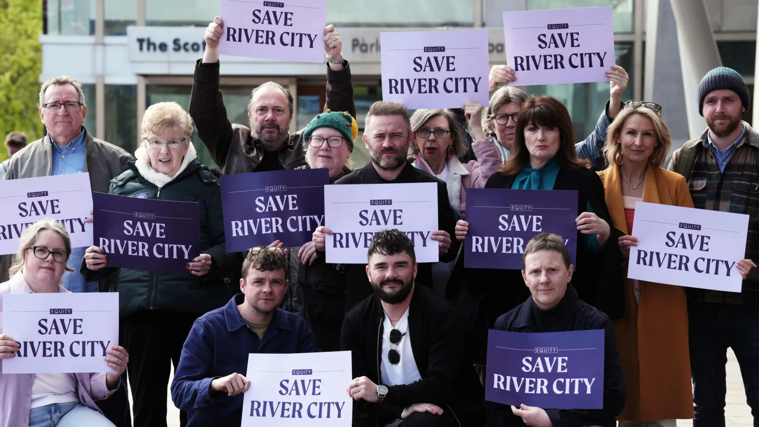  A group of people stand and sit down while outside the Scottish Parliament building. They are all holding placards reading 