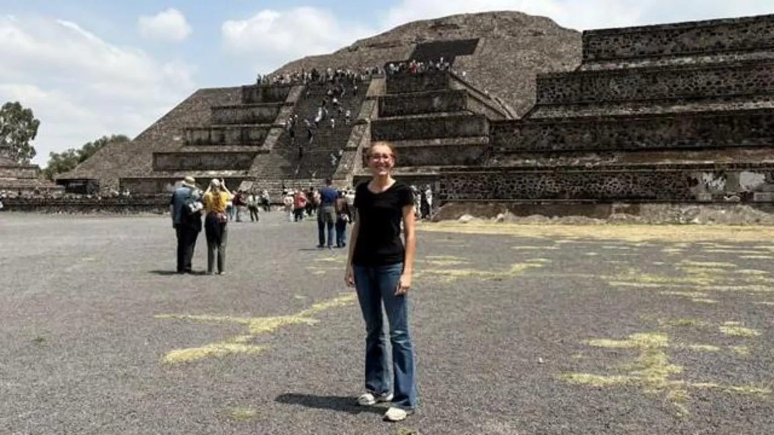 Giovana Guidicini A woman with blue flare jeans and a black t-shirt stands in front of historic monuments scattered with tourists
