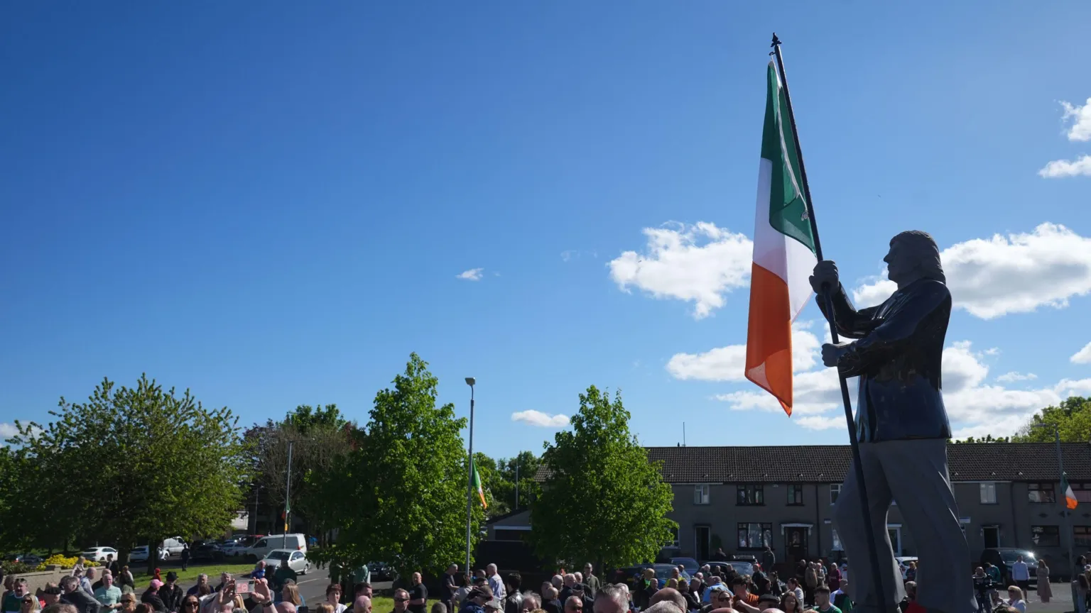  Statue of Bobby Sands in west Belfast. It is a grey statue and there is an Irish tricolour being held up. There is a crowd nearby.