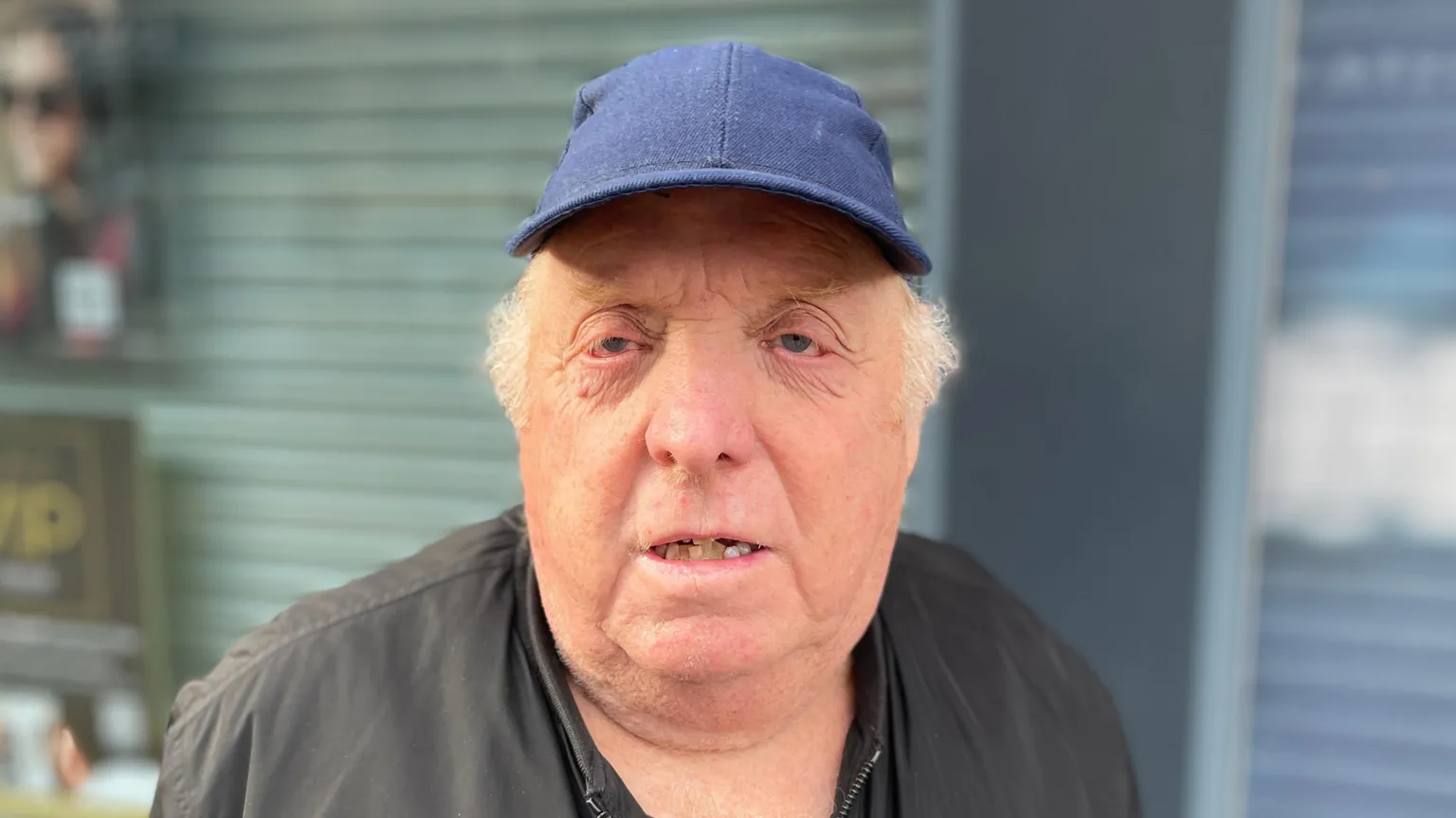 Paul Goodwin, 69, wears a dark blue baseball cap and a black zip-up jacket. He stands outside a shop with a metal shutter partly down. The shop window behind him shows blurred promotional posters. 