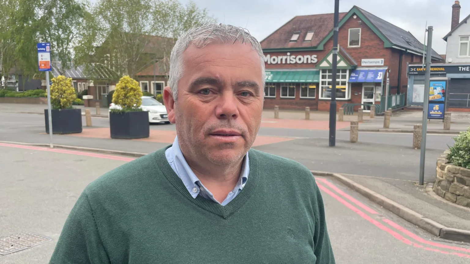 Christian Davis stands on a pavement in Aldridge holding a takeaway coffee cup. He wears a dark green V-neck jumper over a pale blue shirt. The Morrisons store and other shops are visible across the road behind him.