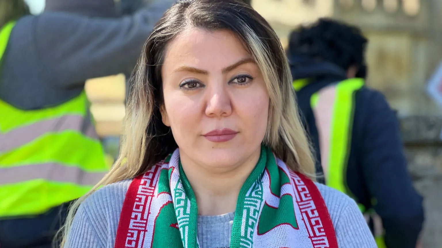 A woman is wearing a scarf in the red, white and green colours of of Iran's flag. She is looking at the camera, with people wearing high visibility jackets walking behind her.