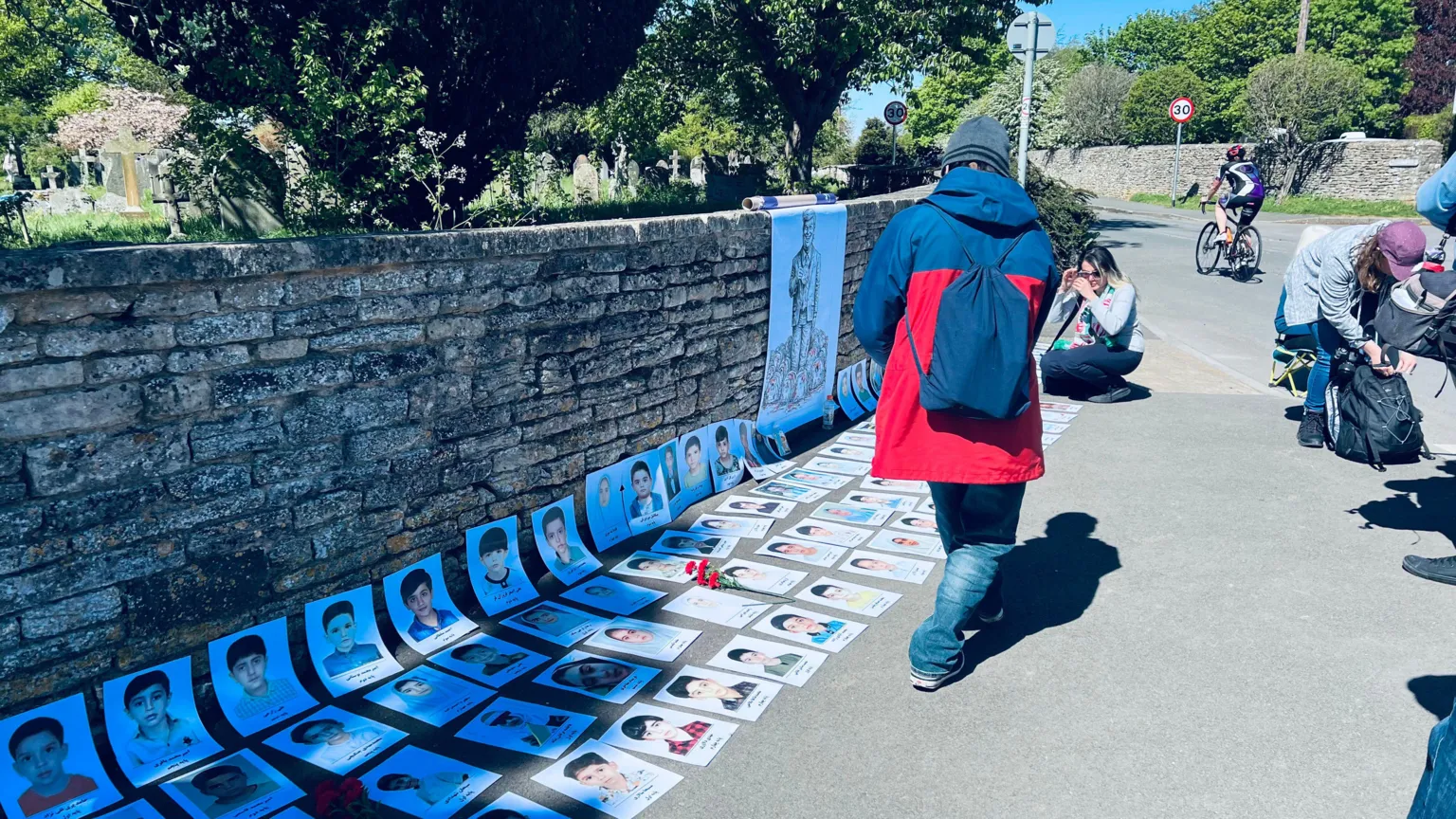 Rows of pictures of children are placed on the floor and balanced against a stone wall. There are people around it.