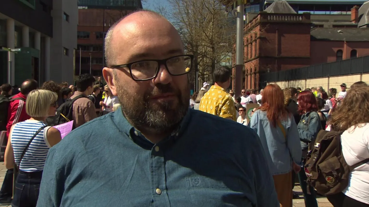 Brendan McNally is wearing black glasses. He's wearing a teal shirt. Crowds of people are behind him in the street. 