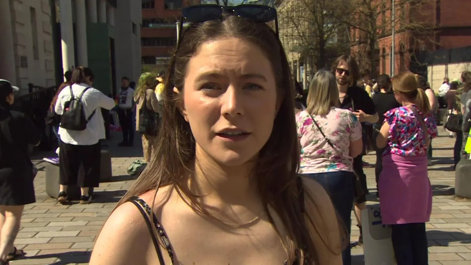 Eva Martin is looking into the camera. She has long brown hair. Sunglasses are on top of her head. People are standing in the background behind her, 