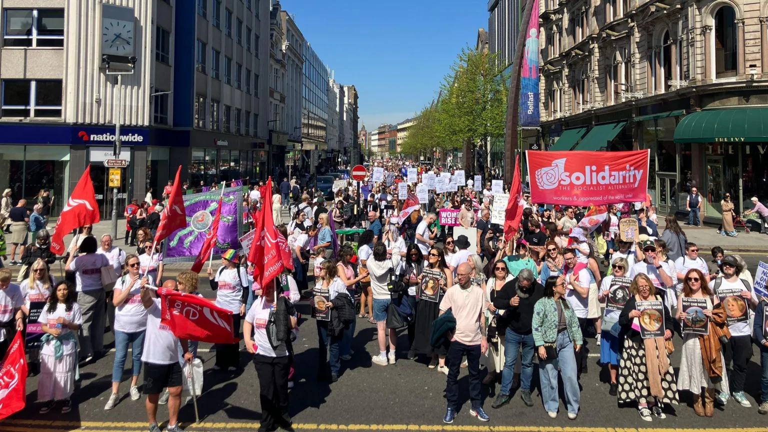 A large crowd are standing on a road. Many are holding banners that says solidarity. Some people are holding photos of women. Shops in a high street are in the background. 