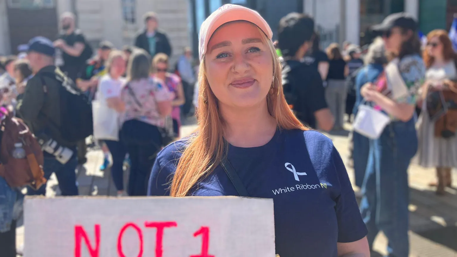 Gillian Lennon is smiling. Her hair is blond. She's wearing a pink cap and a navy top that says White Ribbon NI. Shes holding a banner that says not 1 more. 