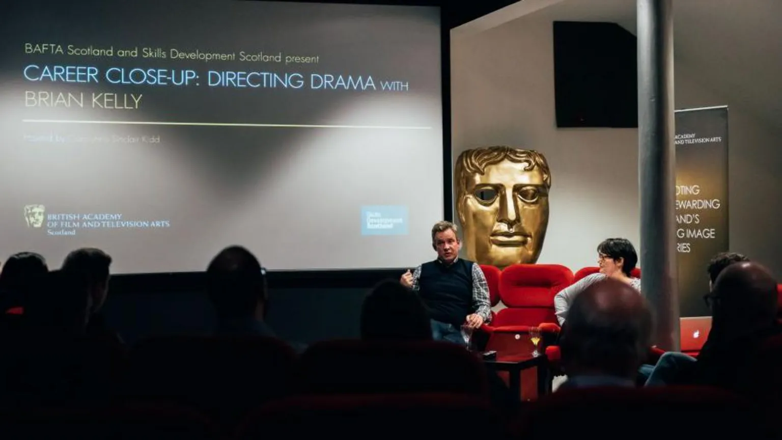  A group of people in a small cinema watching a talk or in conversation event. The big screen shows CAREER CLOSE-UP: DIRECTING DRAMA as the headline.