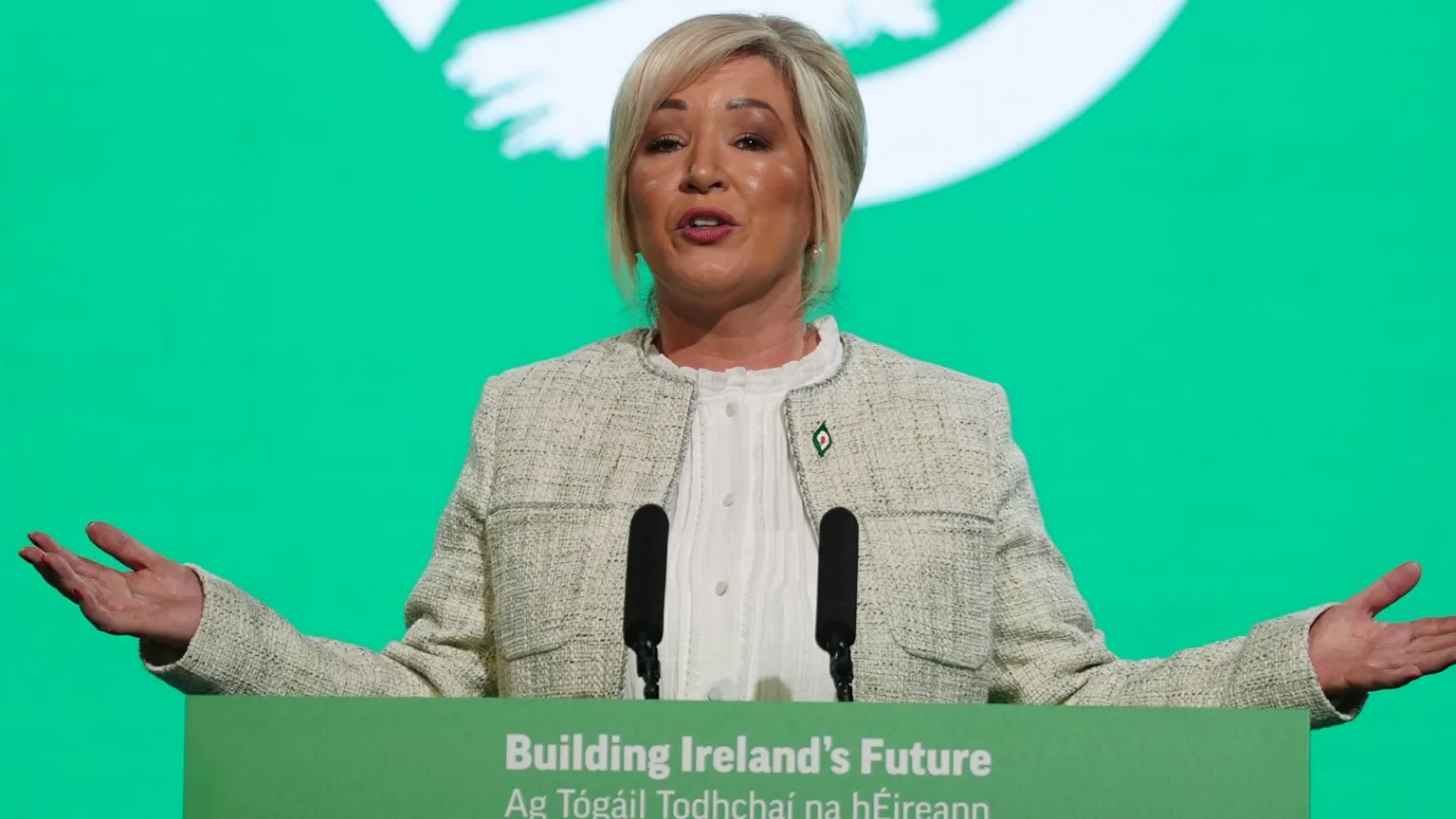  A woman with blonde hair, a grey jacket and white top stands behind a lectern with two black microphones. There is a green and white screen in the background.