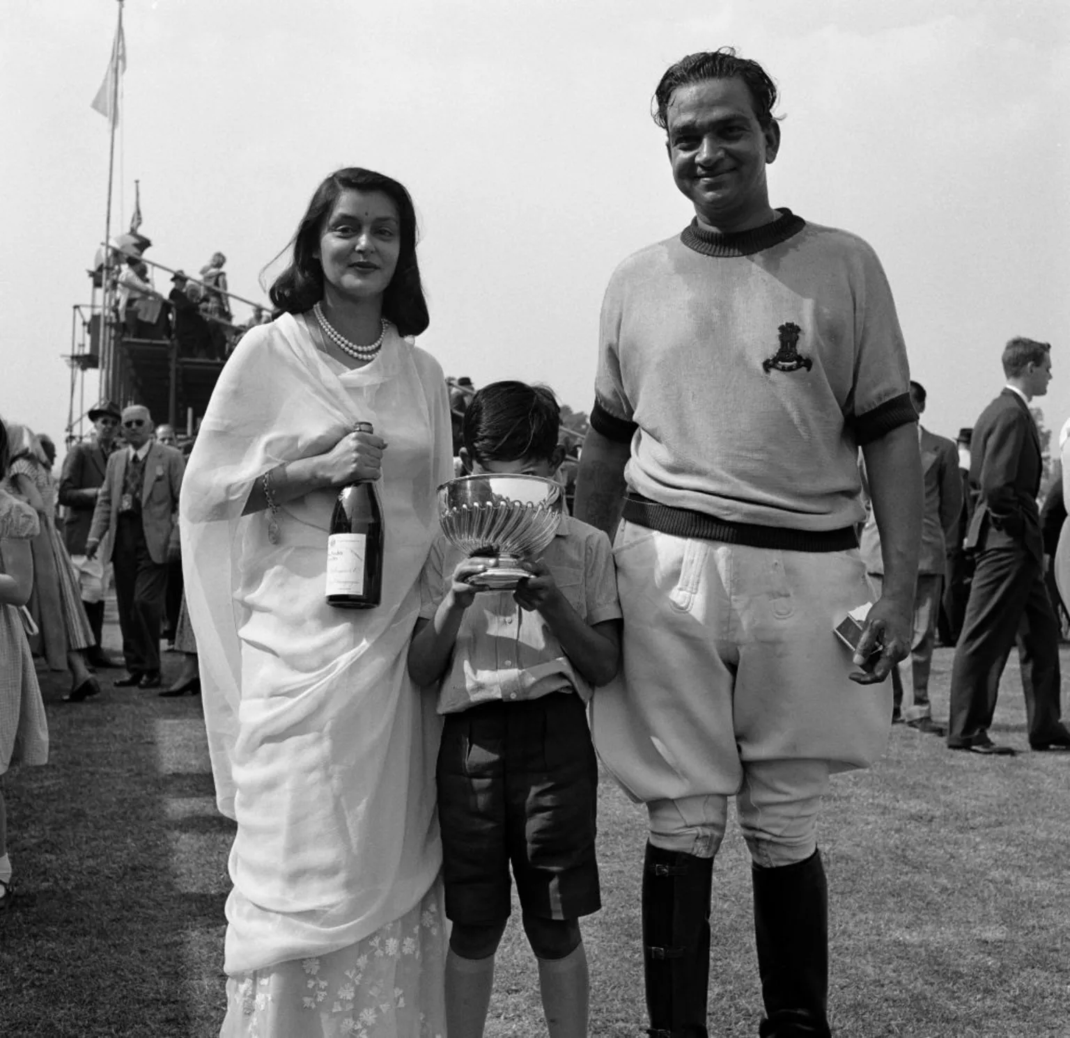  A black and white photo of Sawai Man Singh II (1912 - 1970), the Mahraja of Jaipur, and his wife, Maharani Gayatri Devi (1919 - 2009). The Maharani is holding a bottle of champagne and their son Prince Jagat Singh (1949 - 1997) stands in between, holding a trophy at a polo event in 1957. 