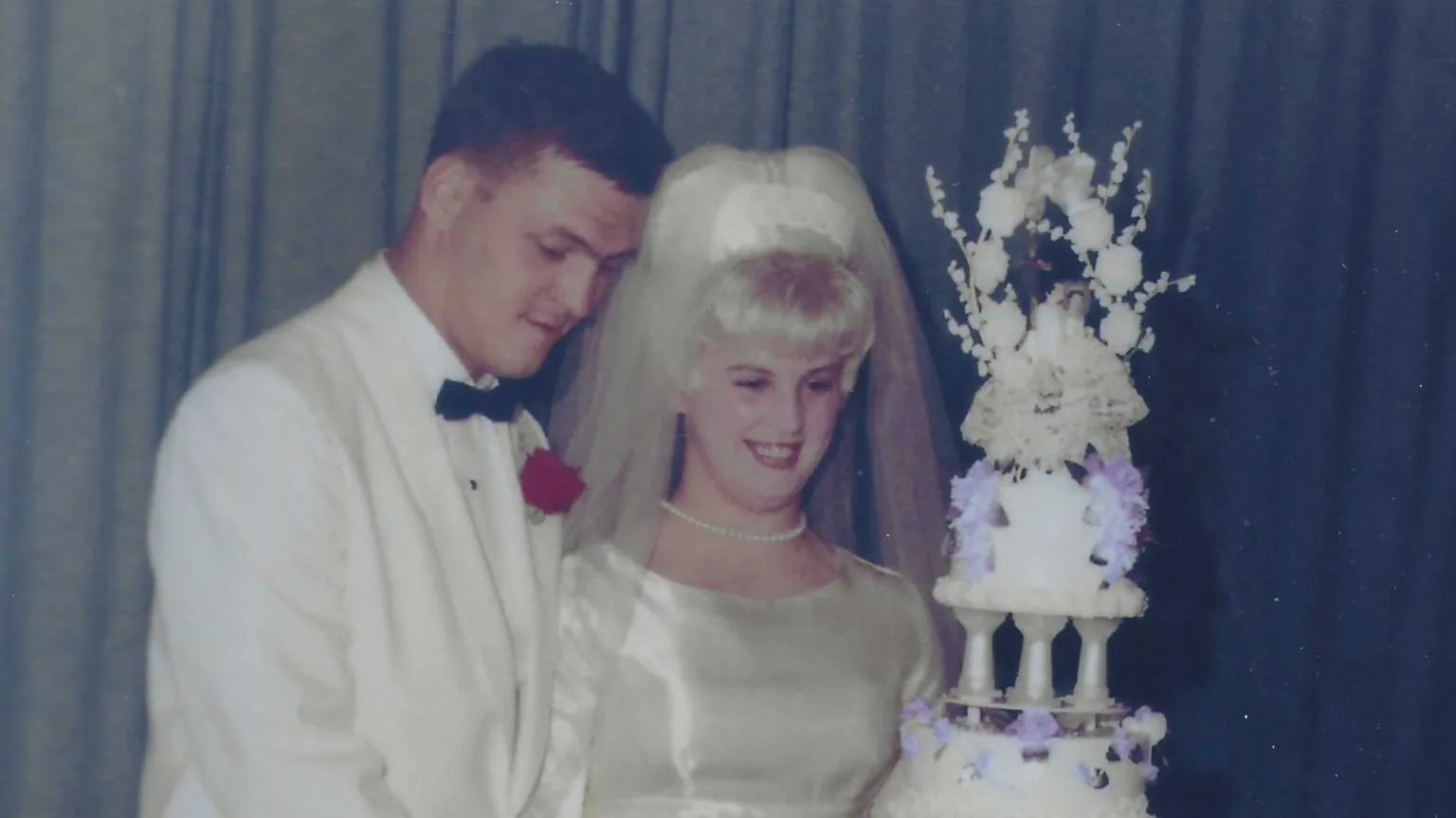 Courtesy of Pam Cronrath An old faded colour photo of a man in a white suit and black bow tie and a smiling woman wearing a white wedding dress and veil. They are standing next to an elaborate multi-tiered wedding cake.