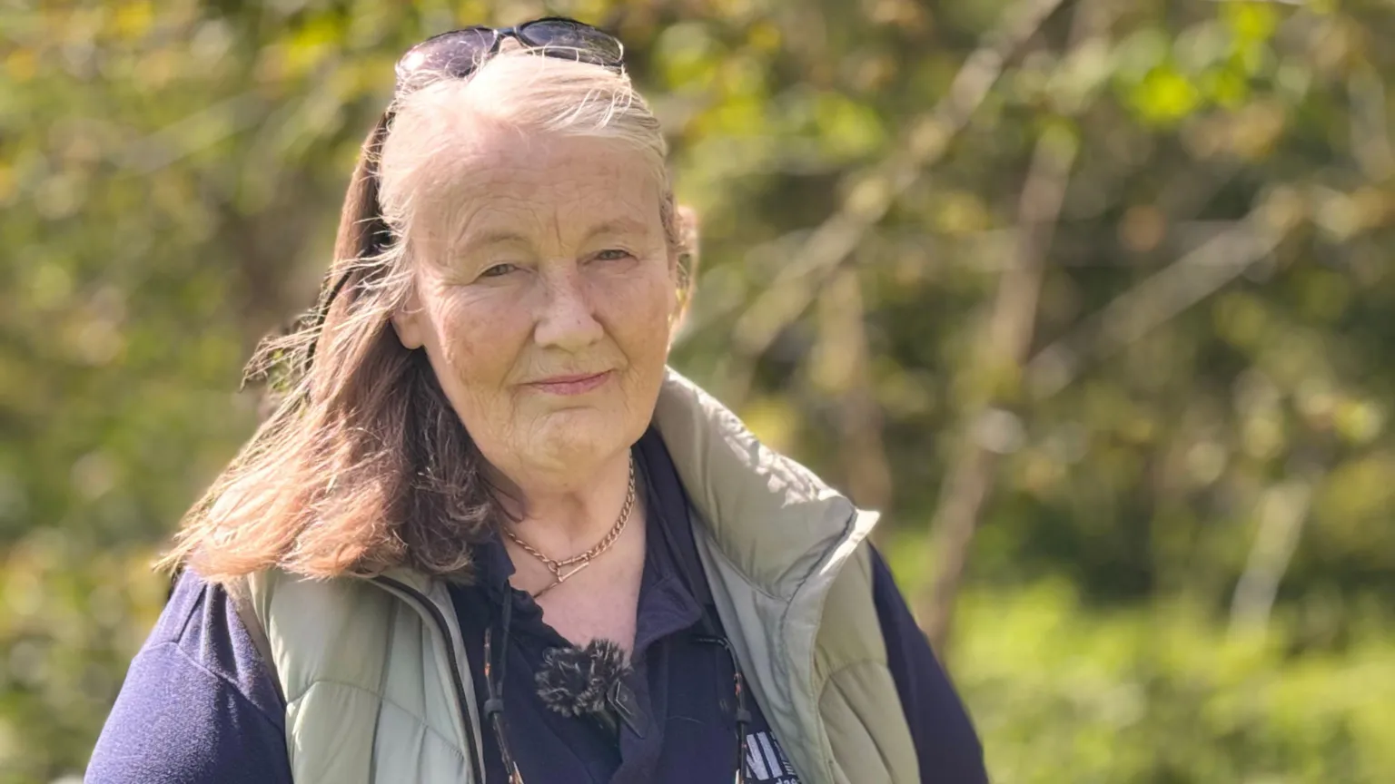 A woman looking at the camera, she has light brown and blonde hair with black sunglasses on top off her head. She is wearing a grey/green gilet and a navy polo shirt.