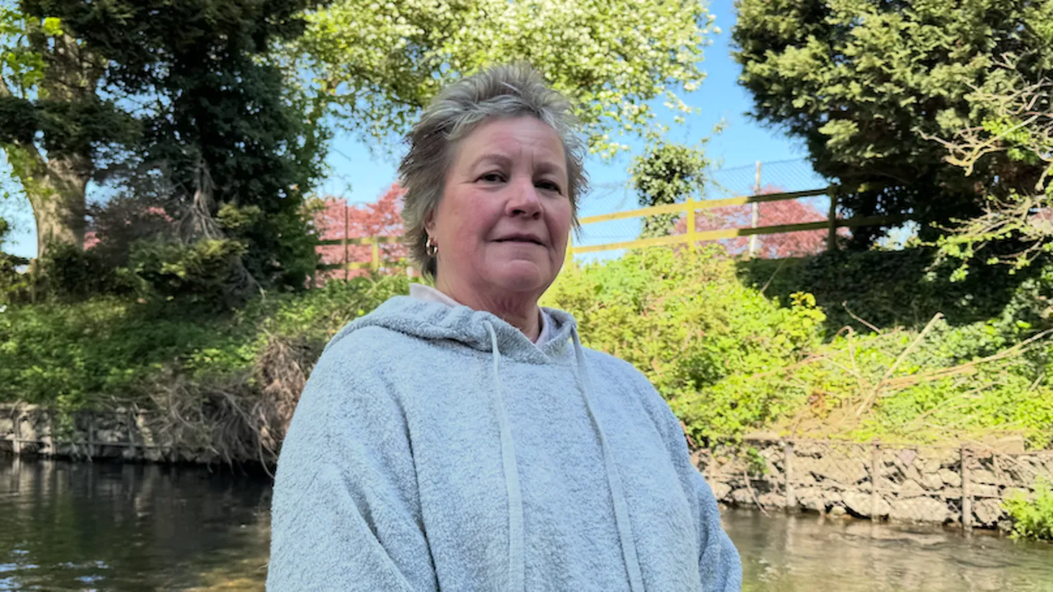 Tessa Smith-Winnard is wearing a grey hooded top and standing in front of a brook. She has short, grey hair. 