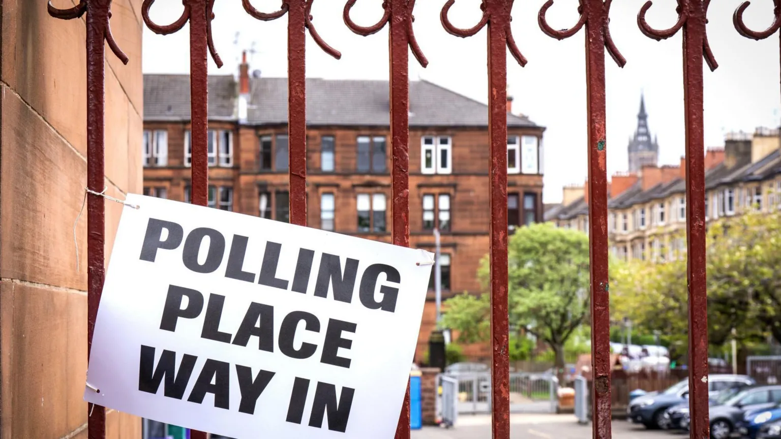  Sign reading “Polling place way in” attached to a red metal gate outside a brick building, with residential buildings and trees visible in the background.