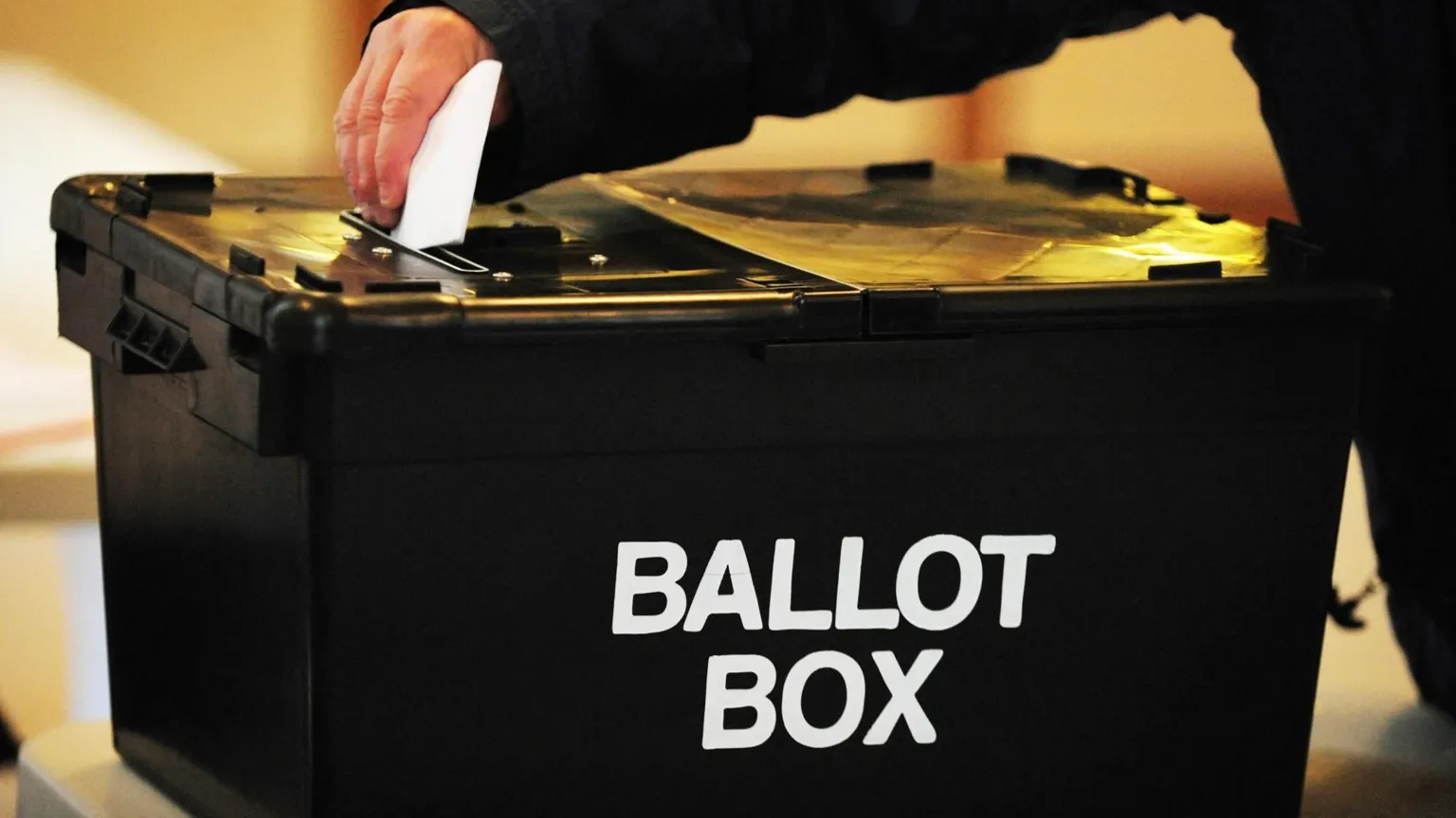  Hand placing a white ballot paper into a black ballot box labeled “BALLOT BOX” inside a polling station.