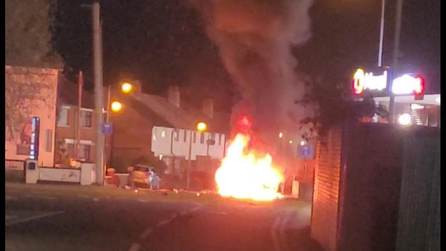 Resident's image At night on a road, a car is on fire near a building. Bright flames and thick black smoke rise into the air. Streetlights and nearby houses can be seen in the background.