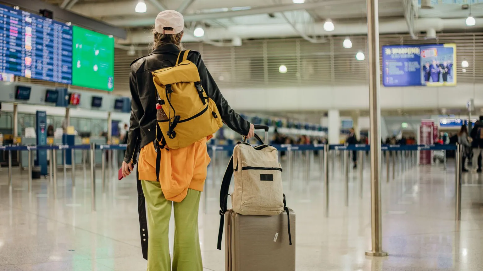  A woman stands in an airport looking at boards showing flights. She is holding a suitcase and backpack.