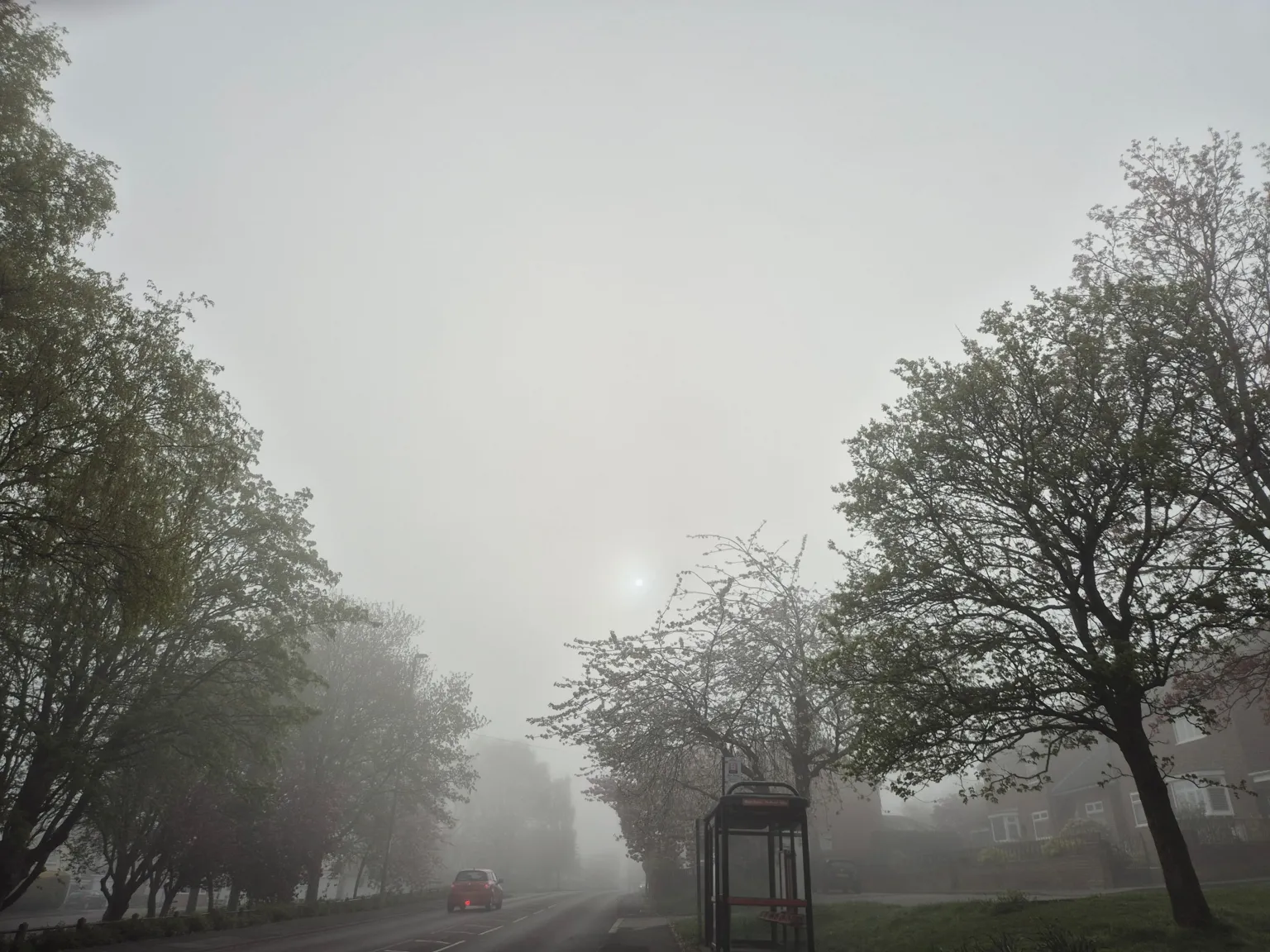 kerry w A foggy road in Ryton, Gateshead, with a car driving away. There is a bus stop on the right and the road is flanked by trees. 