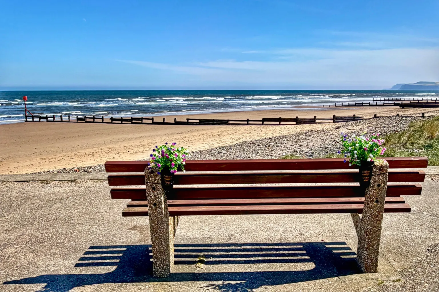Bettys Mum A bench looking over to the sea at Redcar. The bench has two bunches of colourful flowers tied to it. The ky is blue and cloudless, with the beach stretching into the distance. There are no people in view.