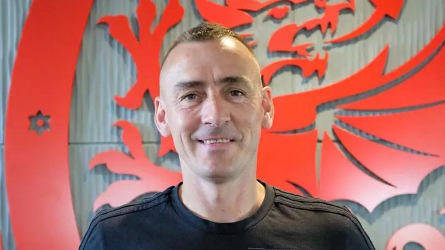 The Homeless World / Street Football Wales Headshot of a middle aged man wearing a black t-shirt with a Welsh dragon logo behind him