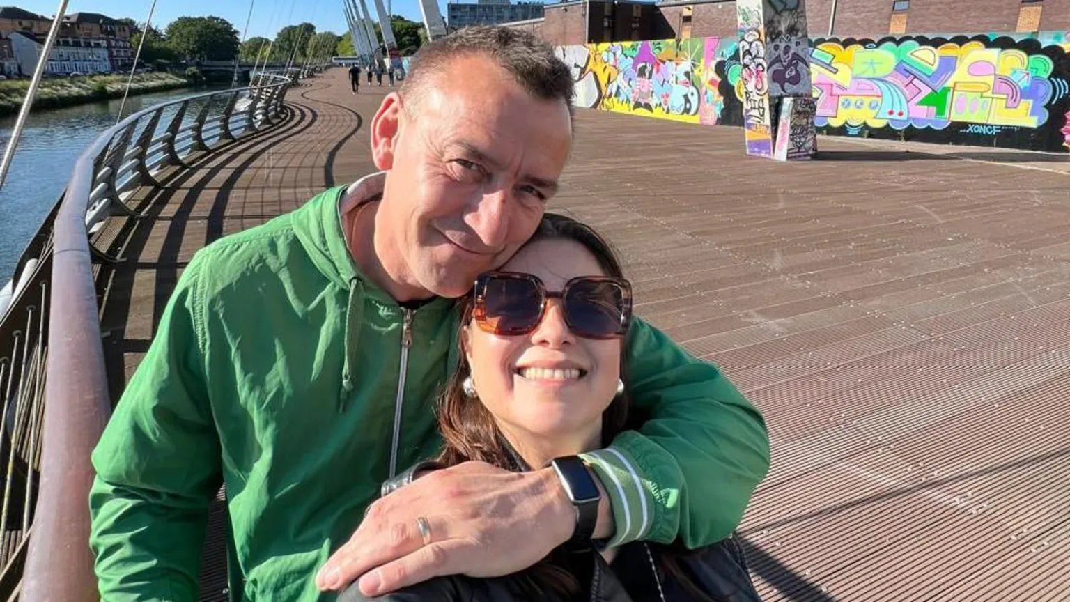 Wayne Ellaway Selfie of a middle aged man and woman smiling, they are on a bridge and it is a sunny day, she is wearing sunglasses he is wearing a green jacket