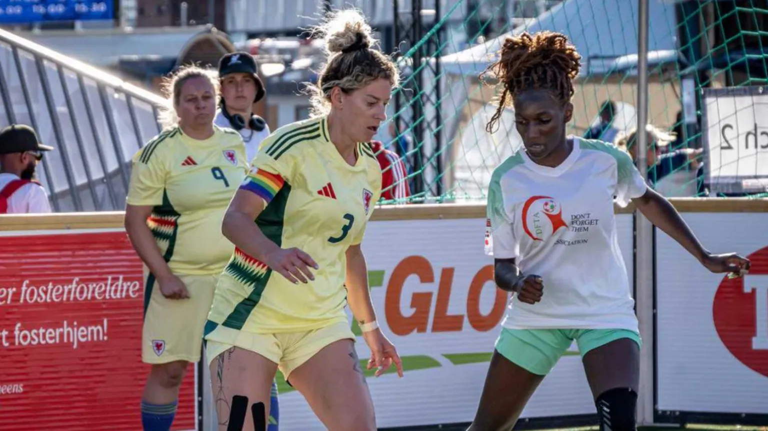 The Homeless World / Street Football Wales There’s a woman playing football in a yellow jersey there is also a woman in a white and pale green uniform.