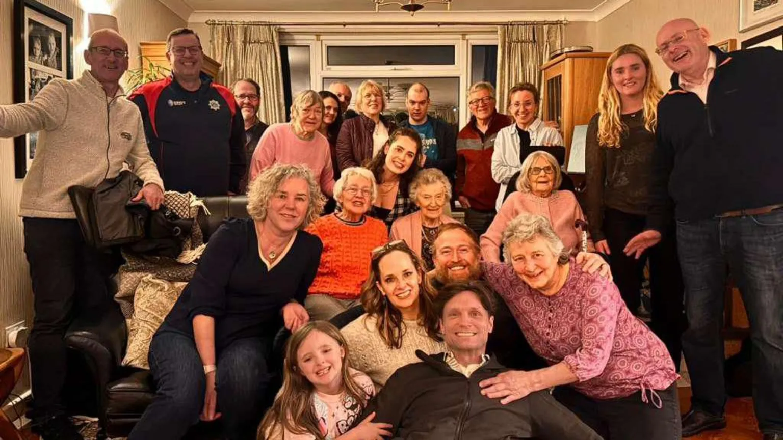 Hans Obma 22 people stand in frame in a living room. Hans Obma is at the centre right at the bottom. Everyone smiles at the camera. 